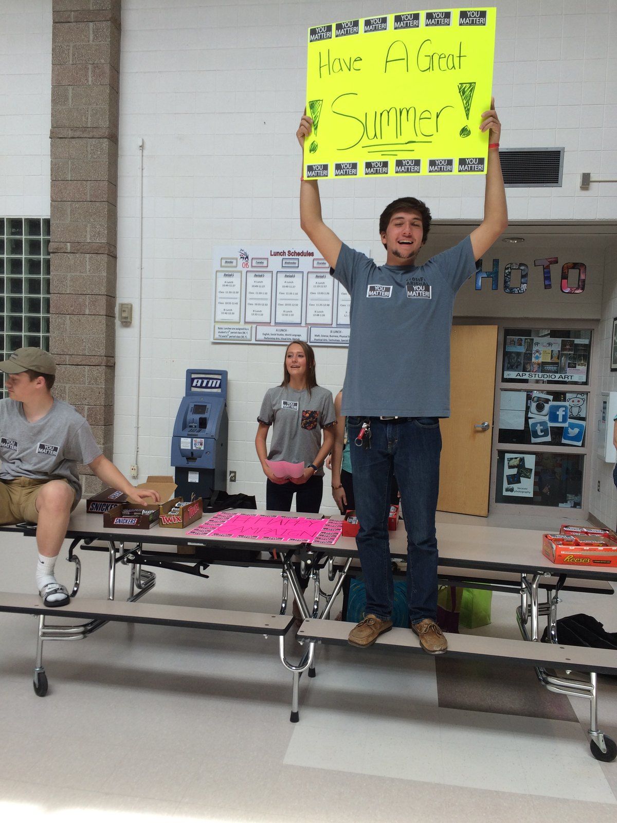 A teen holding up a Have a Great Summer sign covered in You Matter stickers in a school cafeteria, May 2015